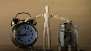 a wooden figure posing one hand on a clock and the other on a money jar a wooden figure posing one hand on a clock and the other on a money jar